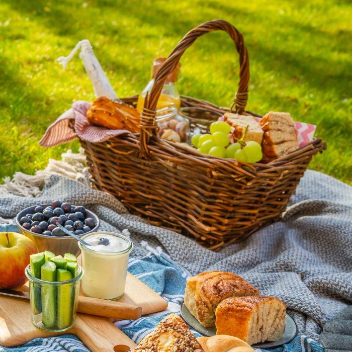 Cesta de piquenique de vime sobre manta no gramado, com uvas, pães, iogurte, frutas e legumes.