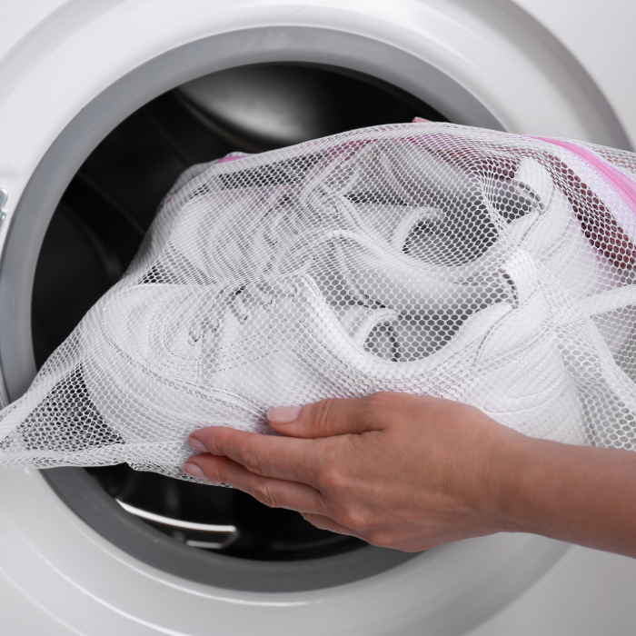 Tênis branco dentro de um saco de lavagem de malha sendo inserido em uma máquina de lavar roupa por uma mão feminina.