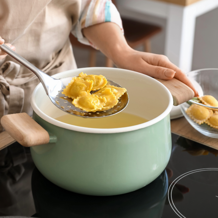 Pessoa segurando uma escumadeira de aço inox com raviólis cozidos sobre uma panela verde clara com alças de madeira, enquanto transfere a massa para uma tigela de vidro ao lado. A cena ocorre em uma cozinha moderna com fogão de indução.