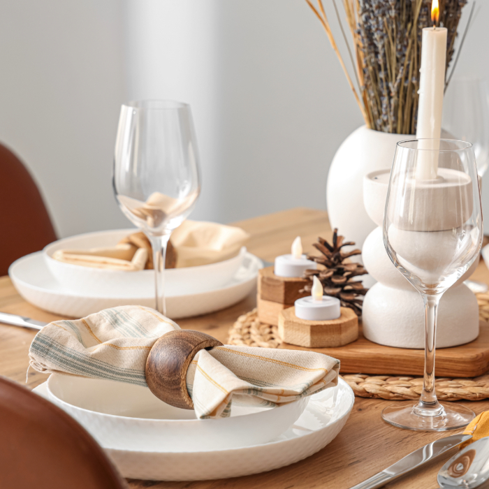 Mesa decorada para jantar com guardanapos dobrados e presos por anéis de madeira, acompanhados de taças de vinho, velas e detalhes rústicos.