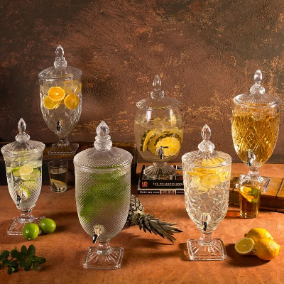 Conjunto de cinco suqueiras de vidro com torneira e tampa decorativa, contendo bebidas com frutas, dispostas sobre mesa de madeira.