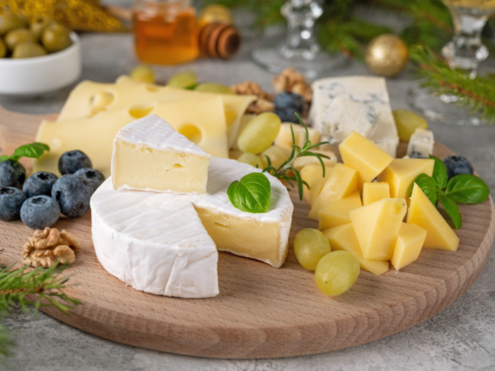 Tábua de petiscos redonda de madeira com divisórias, contendo uma variedade de itens como amêndoas, nozes, fatias de salame, cubos de queijos amarelo e branco, pedaços de queijo brie com nozes e um pequeno recipiente com azeite e um palito decorativo. Ao lado da tábua, há um tecido com estampa colorida, criando uma apresentação rústica e apetitosa.