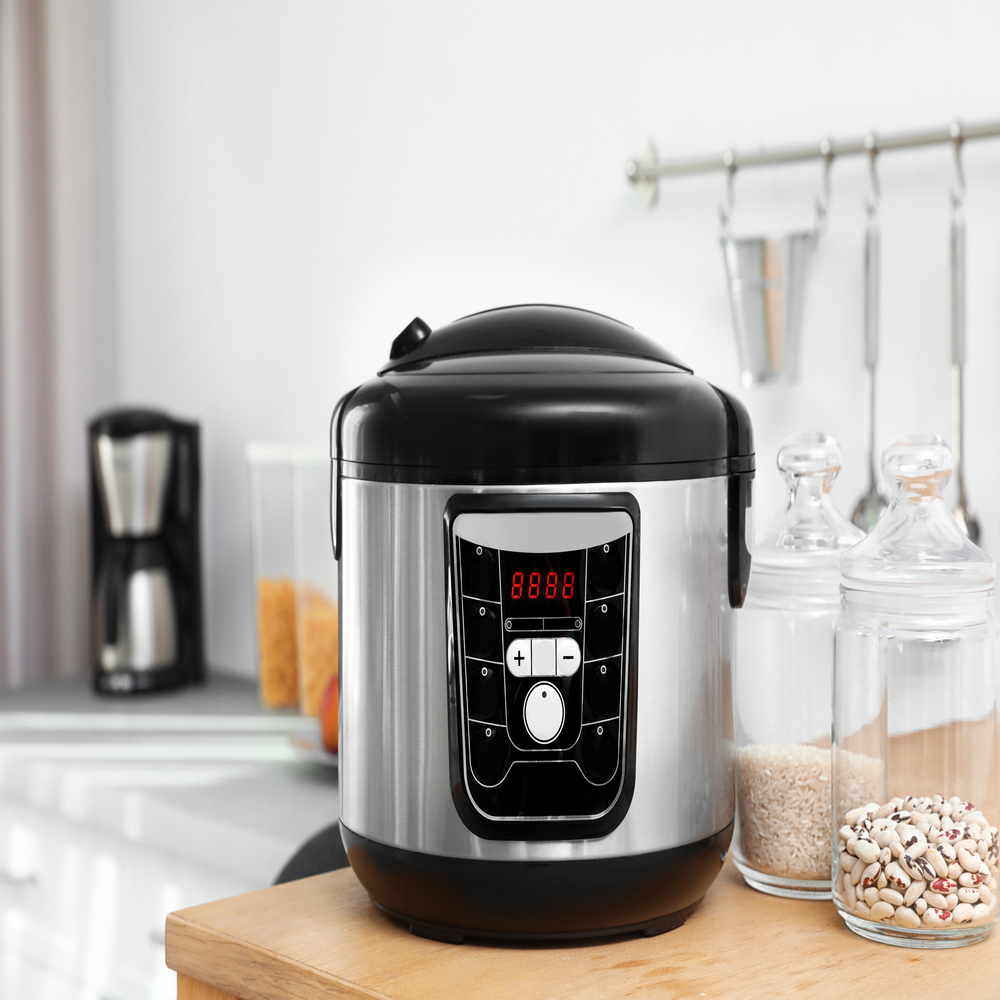 Panela de arroz elétrica em inox e preto posicionada sobre bancada de madeira em cozinha moderna, com potes de mantimentos ao fundo, ideal para preparo de arroz e outras receitas.