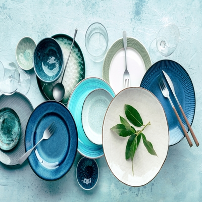 Composição de mesa posta com pratos em tons de azul e branco, taças, talheres e folhas verdes sobre fundo claro, ideal para uma decoração moderna e sofisticada.