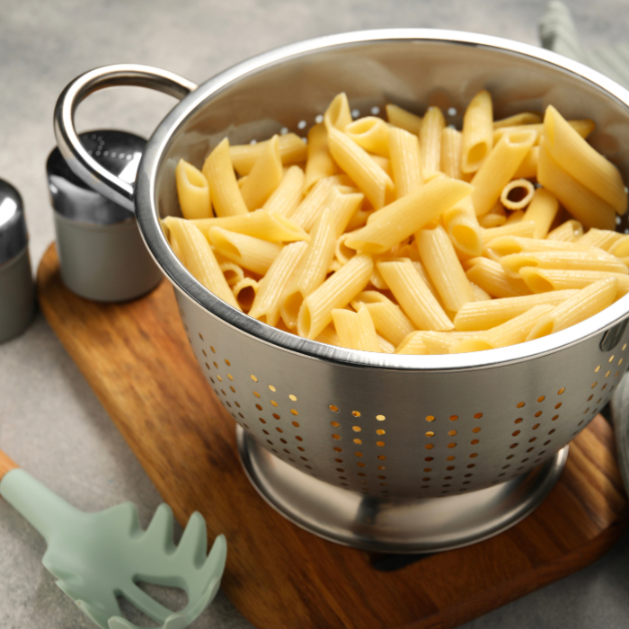Macarrão penne escorrido em escorredor de inox sobre tábua de madeira. Ao lado, utensílio de silicone verde para massa e saleiros metálicos, em uma bancada de cozinha.