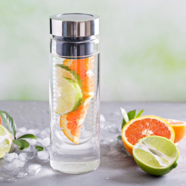 Garrafa infusora de vidro com tampa metálica contendo fatias de laranja e limão, cercada por frutas cítricas e gelo.