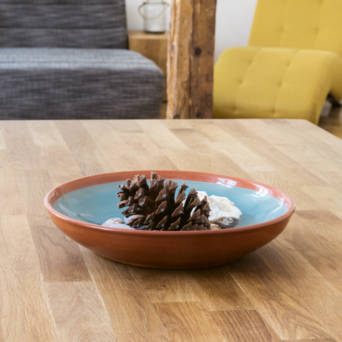 Tigela de cerâmica rústica com exterior terracota e interior azul fosco, colocada sobre uma mesa de madeira. Dentro da tigela, há uma pinha e algumas pedras decorativas. Ao fundo, um ambiente aconchegante com sofá cinza, poltrona amarela e detalhes em madeira, criando uma atmosfera acolhedora e moderna.