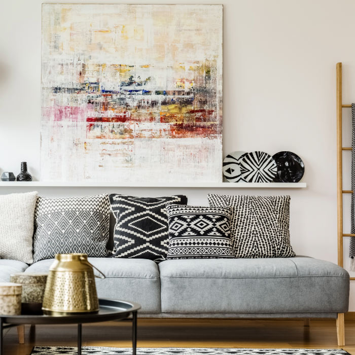 Sala de estar moderna com sofá cinza decorado com almofadas em preto e branco de padrões geométricos, mesa de centro com detalhes dourados, prateleira com objetos decorativos e quadro abstrato na parede.