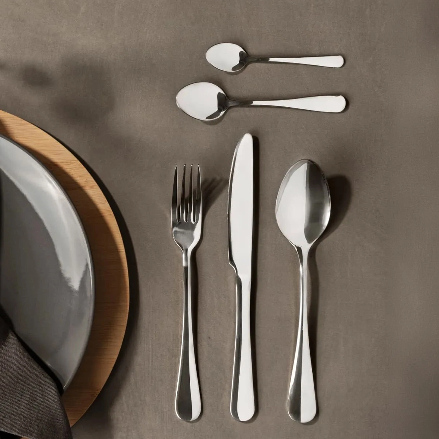 Faqueiros de aço inox com garfo, faca, colher de sopa, colher de sobremesa e colher de chá organizados ao lado de um prato em uma mesa posta minimalista.