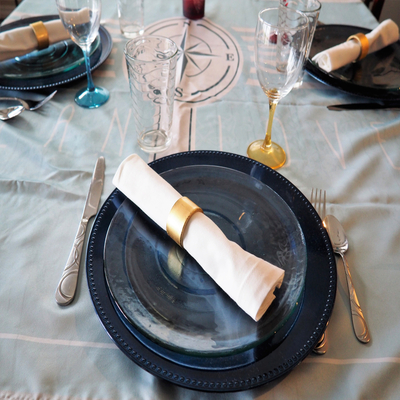 Sousplat azul compondo uma mesa posta sofisticada com prato de vidro, guardanapo branco com anel dourado, talheres e taças, ideal para um jantar elegante.