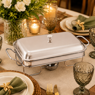 Rechaud retangular de inox com tampa sobre mesa posta decorada com pratos, taças, velas e arranjo de flores.