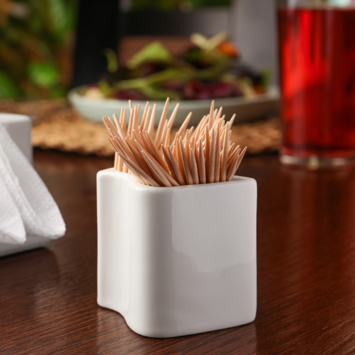 Porta-palitos de dente branco de cerâmica com palitos de madeira, sobre uma mesa de madeira com guardanapos ao lado e uma salada desfocada ao fundo.