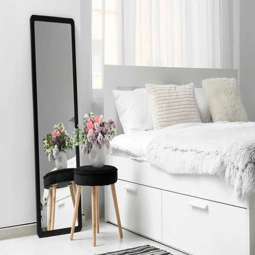 Espelho de corpo inteiro com moldura preta apoiado no chão em quarto moderno, ao lado da cama e de banco decorativo com vasos de flores.