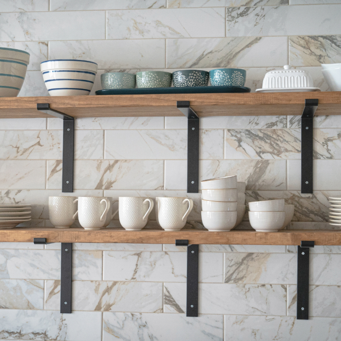 Prateleiras de madeira com suportes pretos, organizadas com xícaras, tigelas e pratos em uma cozinha com parede de azulejos marmorizados.