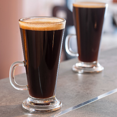 Canecas de vidro com base elevada servindo café preto, dispostas sobre bancada em ambiente de cafeteria.