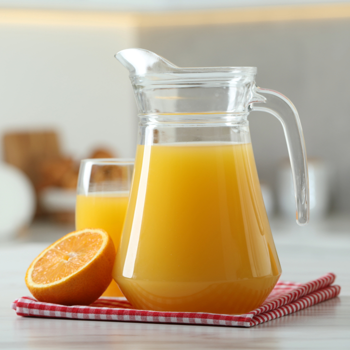 Jarra de vidro transparente cheia de suco de laranja, acompanhada por um copo com suco e uma laranja cortada ao meio, sobre um pano xadrez vermelho e branco em uma mesa clara, com fundo desfocado de cozinha.