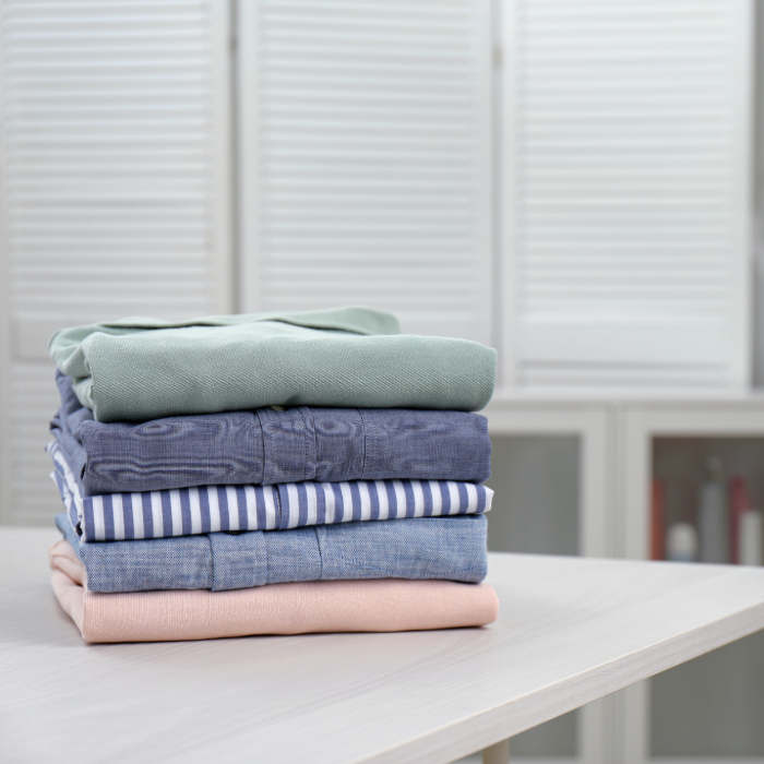 Pilhas de roupas dobradas organizadas sobre uma mesa branca, com armário ao fundo e aparência de lavanderia doméstica.
