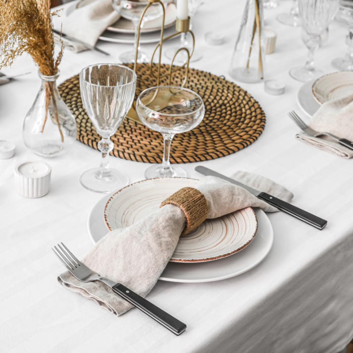 Mesa de jantar elegantemente posta com uma toalha branca, louças de cerâmica com detalhes rústicos, talheres de cabo preto e taças de cristal. O guardanapo de linho é adornado com um anel de fibra natural. No centro da mesa, há um sousplat trançado, velas decorativas e arranjos secos em vasos de vidro, criando uma atmosfera sofisticada e acolhedora.