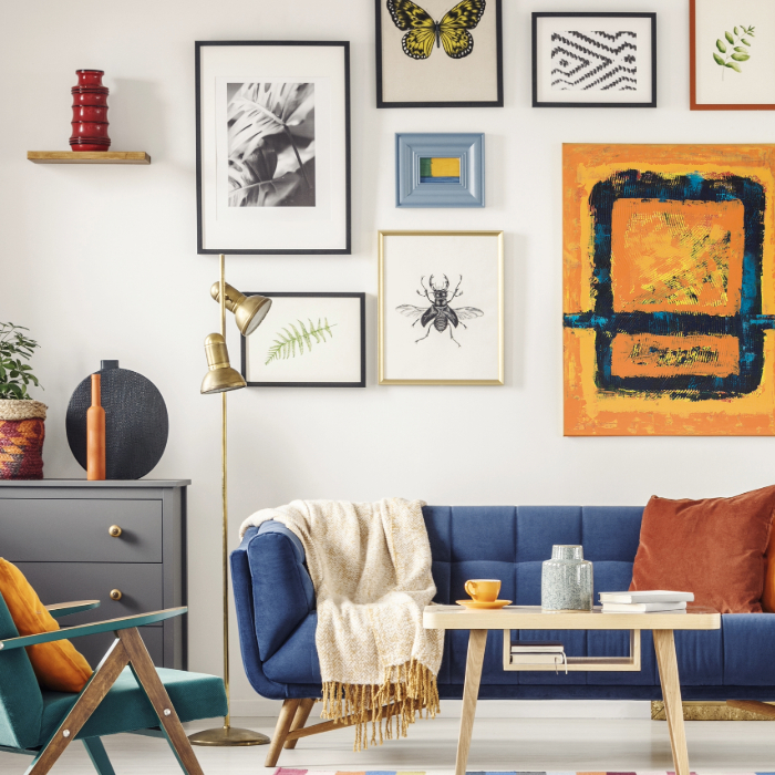 Sala de estar colorida com sofá azul, poltrona verde, almofadas laranjas, mesa de centro de madeira e parede branca decorada com quadros variados, incluindo arte abstrata e ilustrações botânicas.