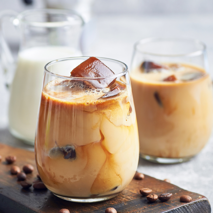 Copos de vidro com café gelado e cubos de gelo de café, acompanhados por uma jarra de leite ao fundo e grãos de café espalhados sobre a superfície de madeira.