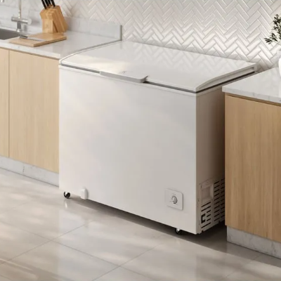 Freezer horizontal branco com tampa superior e controle de temperatura frontal, posicionado entre armários de madeira clara em cozinha planejada.