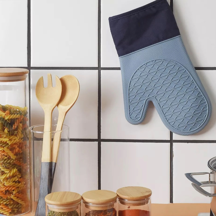 Luva térmica azul pendurada em parede de azulejos brancos, ao lado de utensílios de cozinha e potes com massas e temperos.