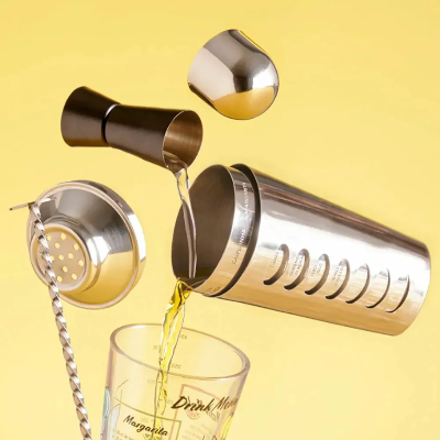 Kit de acessórios para drinks em aço inox, com copo dosador duplo, coqueteleira, peneira e colher bailarina, exibidos sobre fundo amarelo com bebida sendo servida em copo medidor.