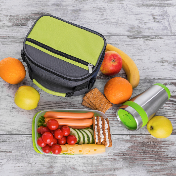 Bolsa térmica cinza e verde com lanche saudável ao lado, incluindo salsichas, queijo, tomate-cereja, pepino fatiado, biscoitos e frutas como banana, maçã e laranja.
