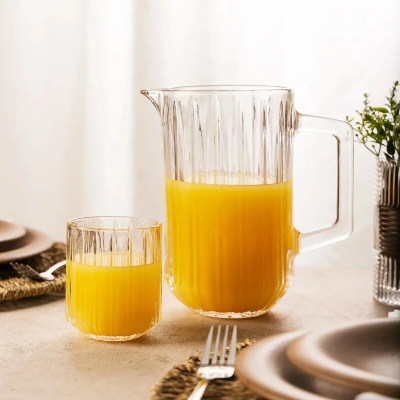 Jarra de vidro canelado com alça e bico, contendo suco de laranja, acompanhada de copo sobre mesa posta.
