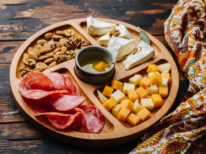 Tábua de petiscos redonda de madeira com divisórias, contendo uma variedade de itens como amêndoas, nozes, fatias de salame, cubos de queijos amarelo e branco, pedaços de queijo brie com nozes e um pequeno recipiente com azeite e um palito decorativo. Ao lado da tábua, há um tecido com estampa colorida, criando uma apresentação rústica e apetitosa.