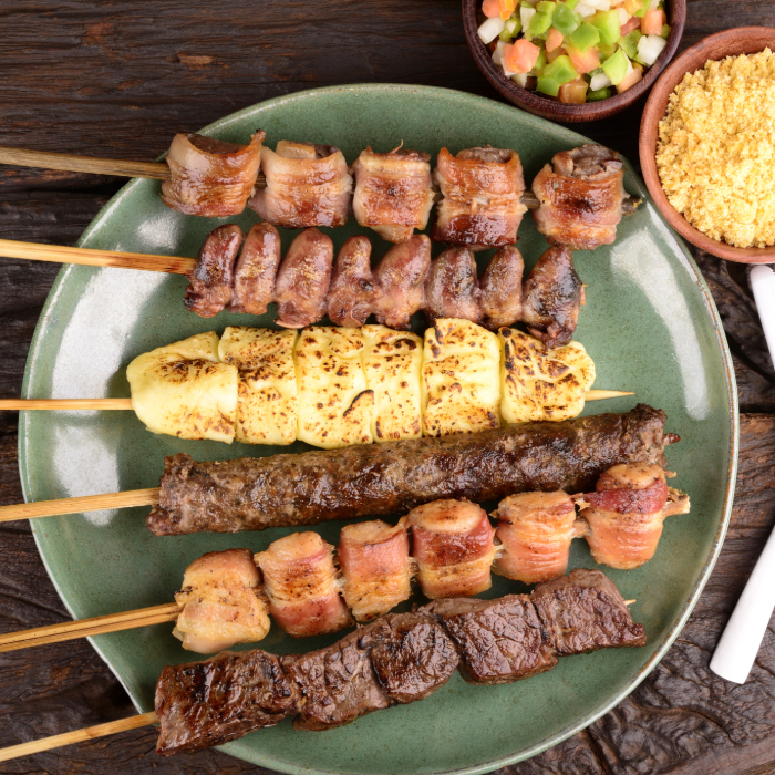 Prato verde com diversos espetinhos de churrasco, incluindo carne, frango, queijo coalho, coração de frango e bacon, acompanhado de farofa e vinagrete em potes de barro sobre uma mesa rústica.