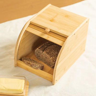 Porta-pão de bambu com tampa deslizante e puxador redondo, contendo pão integral fatiado, sobre uma mesa com toalha clara.