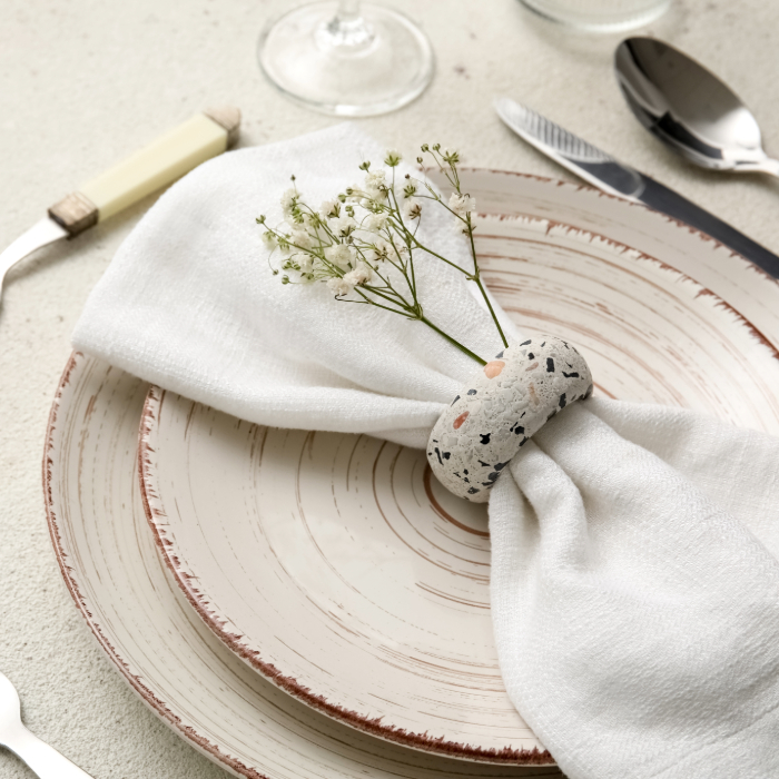 Mesa posta com guardanapo branco dobrado e preso por um anel de guardanapo decorativo com pequenas flores brancas, sobre pratos rústicos e talheres elegantes.