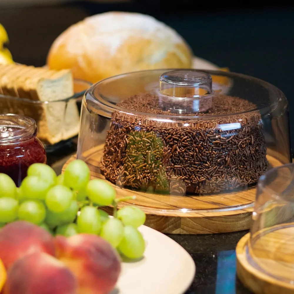 Boleira redonda com base de madeira e tampa transparente, exibindo bolo de chocolate com granulado sobre uma mesa com frutas, pães e geleia.