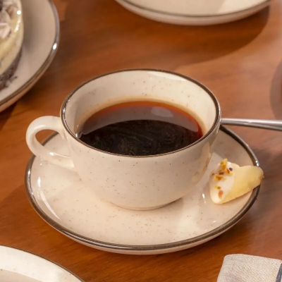 Xícara de cerâmica bege com acabamento pontilhado e borda escura, servida com café preto sobre pires combinando em mesa de madeira.