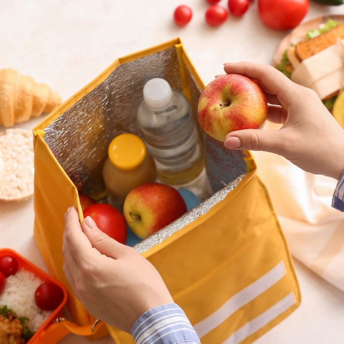 Pessoa pegando uma maçã dentro de uma bolsa térmica amarela com isolamento interno prateado, contendo garrafa de água, suco e alimentos frescos.