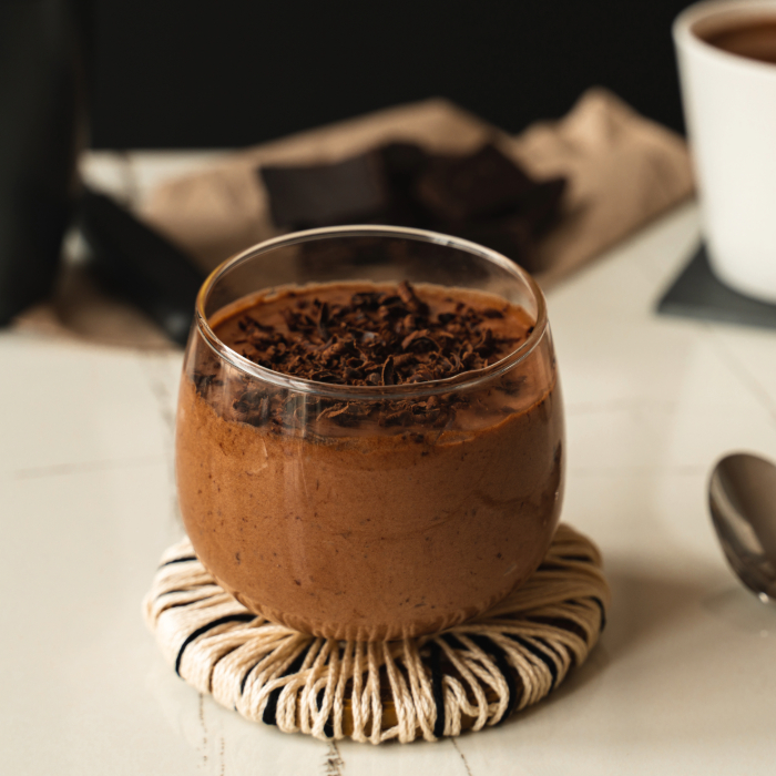 Taça de vidro com mousse de chocolate decorado com raspas de chocolate por cima, apoiada em descanso de corda trançada sobre uma mesa branca. Ao fundo, pedaços de chocolate e uma xícara branca com bebida quente.
