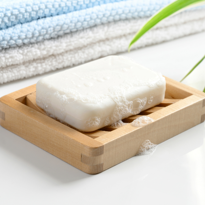 Sabonete branco com espuma apoiado em um porta-sabonete de madeira, com toalhas dobradas ao fundo.