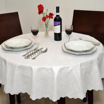 Toalha de mesa redonda branca adamascada em mesa posta com pratos, talheres, taças e arranjo de flores vermelhas.