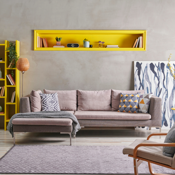 Sala de estar moderna com sofá cinza e almofadas geométricas, nicho amarelo embutido na parede com decoração, estante amarela com livros e plantas, e quadro abstrato apoiado no chão.