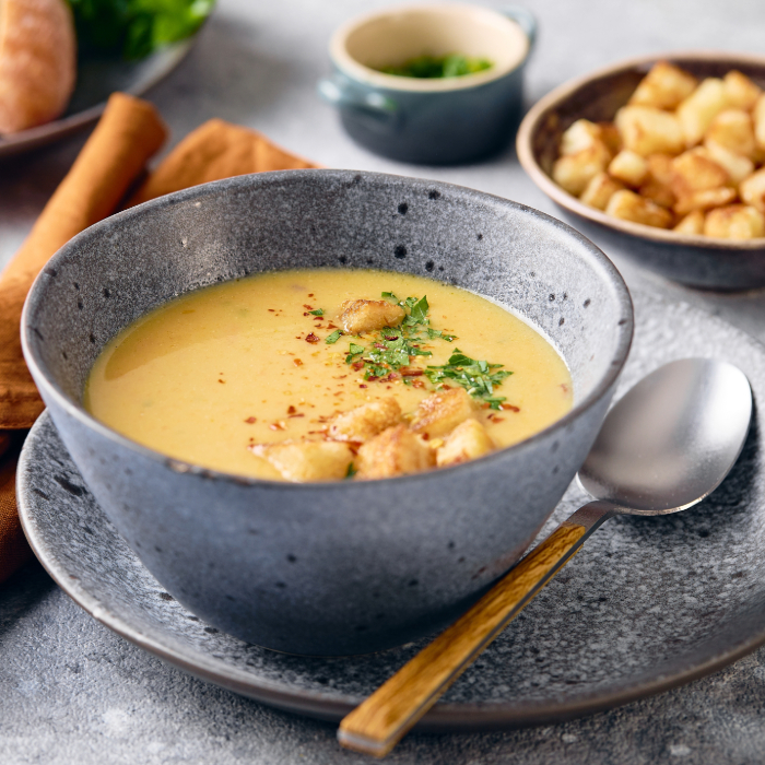 Tigela de sopa cremosa servida com croutons e ervas frescas, acompanhada de colher sobre um prato cinza.