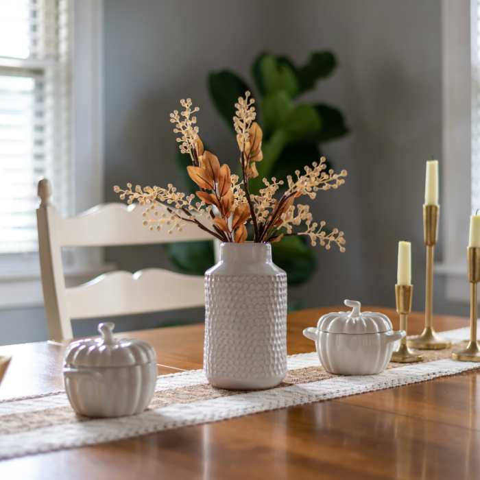 Mesa de jantar decorada com um vaso de cerâmica branca texturizada contendo arranjo de folhagens secas em tons terrosos. Ao lado, há dois potes decorativos em formato de abóbora branca e castiçais dourados com velas. A mesa tem um caminho de mesa de tecido rendado e, ao fundo, cadeiras brancas e uma planta verde em um ambiente bem iluminado.