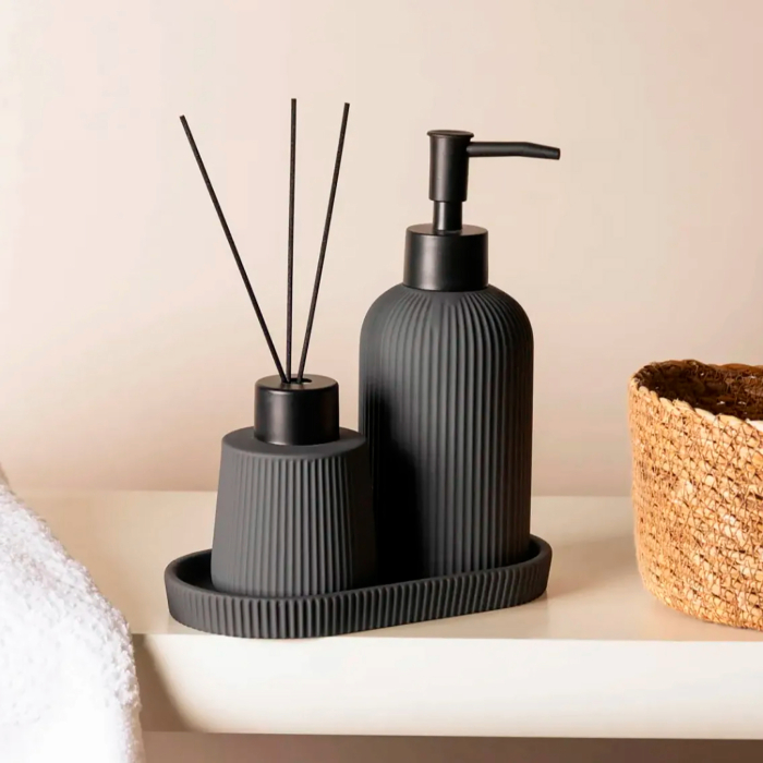 Conjunto de banheiro moderno com acabamento preto fosco e textura em relevo, composto por aromatizador com varetas e dispenser de sabão líquido sobre bandeja oval, posicionado em bancada clara com cesto de palha ao lado.