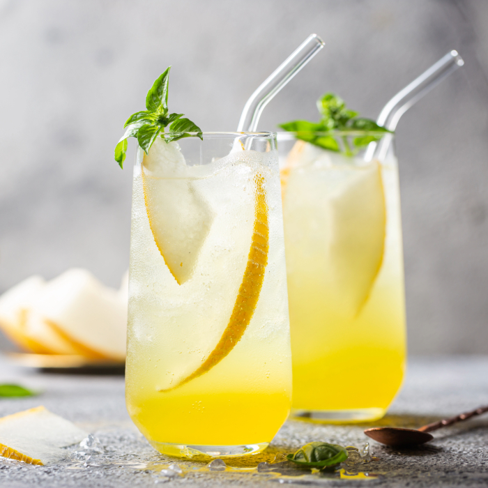 Duas bebidas refrescantes amarelas servidas em copos altos com canudos de vidro, decoradas com fatias de limão, pedaços de melão e folhas de manjericão, sobre uma superfície rústica com gelo derretido.
