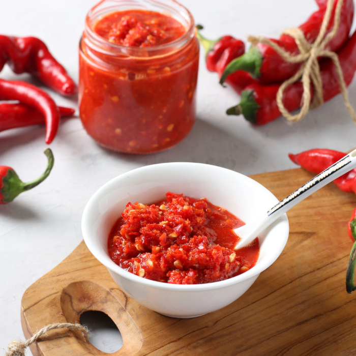 Molho de pimenta vermelha em um pote de vidro e servido em uma tigela branca com colher, rodeado por pimentas frescas.