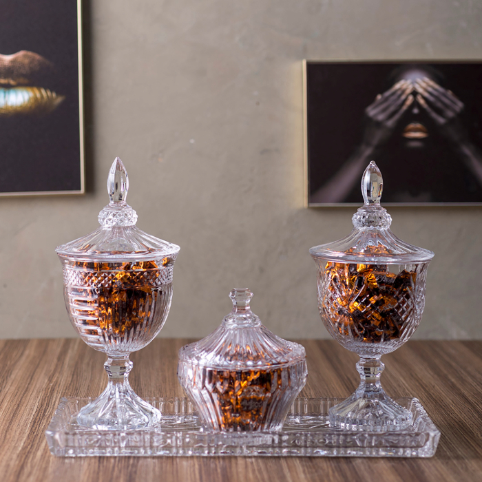 Conjunto refinado de bombonieres de cristal com tampas, apoiadas sobre uma bandeja de vidro em uma mesa de madeira. As peças possuem design sofisticado e estão cheias de bombons embrulhados em papel dourado, criando um toque de requinte à decoração.