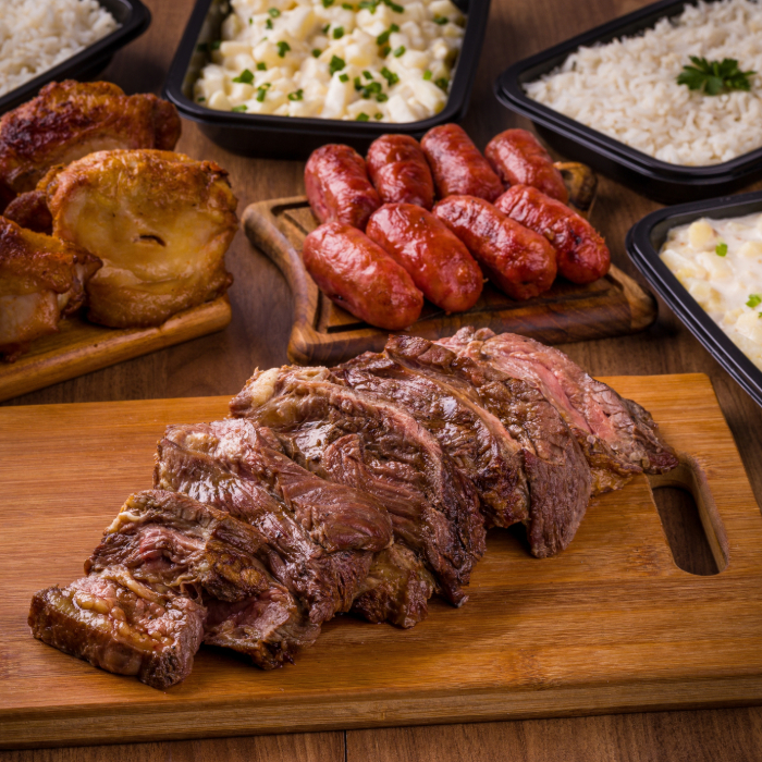 Churrasco servido em tábuas de madeira, com cortes de carne fatiada, linguiças, e acompanhamentos como arroz, maionese e batatas, dispostos sobre uma mesa rústica.