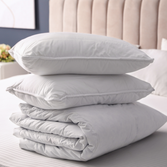 Dois travesseiros brancos empilhados sobre um edredom dobrado, todos sobre uma cama com cabeceira estofada bege e decoração sofisticada.