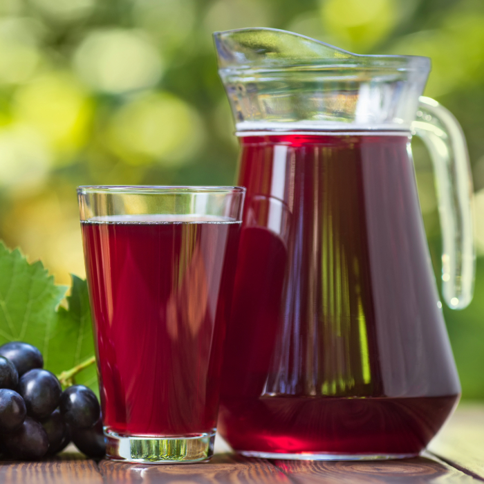 Jarra de vidro transparente cheia de suco de uva ao lado de um copo servido, com cachos de uvas escuras e folhas verdes ao fundo, em uma mesa de madeira ao ar livre.
