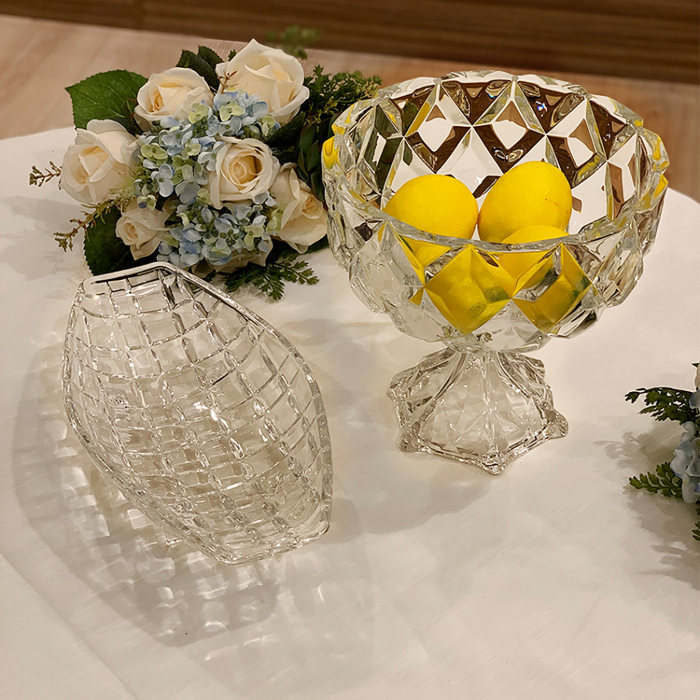 Centro de mesa decorado com uma fruteira de cristal lapidado com pedestal, contendo limões amarelos, ao lado de uma tampa de cristal com textura quadriculada. O cenário é complementado por arranjos florais com rosas brancas e hortênsias azuis, criando um ambiente elegante e sofisticado sobre uma mesa branca.
